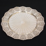 Bandeja circular confeccionada em metal prateado,repuxado com raios incisos convergentes ao centro, orla vazada com reservas de motivois florais, borda ondulada e recortada. Medida: Diam.: