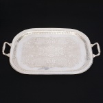 Grande bandeja confeccionada em metal espessurado à prata, formato retangular de quinas arredondadas, borda elevada, revirada para fora e percorrida por faixa relevada, orla circundada por com sucessão de gomos verticais, obtidos através da técnica do repuxado; centro rebaixado, profusamente lavrado com composição de motivo vegetalista, alças laterais cinzeladas com reserva oblonga, frisos e terminações em folhagens. Medidas: Alt.: 5 x Comp.: 66 x Larg.: 42,5 cm.