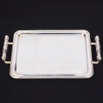 Elegante bandeja confeccionada em metal espessurado à prata, formato retangular de quinas arredondadas, borda percorrida por contorno relevado, centro ligeiramente rebaixado, ricamente lavrado com composição de motivoi vegetalista, alças laterais fusiformes, relevada com folhagens, apoia sobre hastes curvadas. Medidas: Alt.: 5 x Comp.: 40 x Larg.: 26 cm.