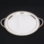 EBERLE. Belíssima bandeja circular confeccionada em metal espessurado à prata, estilo neoclássico, centro ligeiramente rebaixado, borda contornada por barrado relevado e cinzelado com fileira de pétalas, alças laterais em arcos terminadas em folhas de acanto. Marca da manufatura no verso. Med.: Comp(de uma alça a outra).: 46 cm; Diam.: 38 cm.