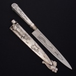 Punhal de coleção confeccionado em metal espessurado à prata, bainha e cabo relevados com cena campestre contendo paisagem, personagens e animais, ponta decorada com porongo, chapéu e boleadeira, lâmina em aço inox da manufatura Elmo. Med.: Comp. 29 cm. Apresenta desgastes no banho de prata.