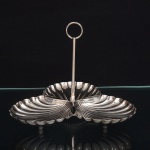 Elegante petisqueira confeccionada em metal espessurado à prata, dotada de três recipientes moldados em forma de concha, apoiados sobre três pés esféricos, interior ligeiramente côncavo, repuxado com sulcos longitudinais, borda recortada em ondulações regulares; haste central tubular encimada por alça circular. Medidas: Alt.: 14,5 x Diam.: 17 cm.