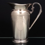 EBERLE. Elegante jarra confeccionada em metal espessurado à prata, bojo balaústre ricamente lavrada com elementos vegetalistas, assente sobre base circular repuxada em degraus, asa lateral em volutas, boda ondulada e revirada para fora, interior dotado de barra retentora de gelo. Marca da manufatura ao fundo. Med.: Alt. 21,5 x Comp. 20 x Larg. 13 cm.