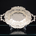 Belíssima travessa oval, estilo rococó, corpo repuxado, aba recortada e moldada, decorada em relevo contínuo de volutas, rocalhas, concheados e cartelas, com reserva central oval lisa. Borda  com recortes simétricos integrados à decoração. Duas alças laterais aplicadas e fundidas, de perfil curvo e seção vazada, decoradas em consonância com a aba. Base apoiada sobre quatro pés fundidos, integrados ao corpo, com terminação decorativa. Peça executada por técnicas combinadas de repuxo, cinzelagem e fundição. Medidas: Alt.: 9 x Comp.: 38 x Larg.: 24,5 cm.
