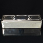 Caixa para conjunto de toucador confeccionada em metal espessurado à prata, formato retangular, tampa removível, superfície decorada com reserva central composta por coroa de folhagens, emoldurada por fileira de frisos intercalados por fitas cruzadas. Medidas: Alt.: 5 x Comp.: 23 x Larg.: 8 cm.