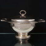 FRACALANZA. Belíssima sopeira confeccionada em metal espessurado à prata, bojo hemisférico assente sobre base circular elevada, tampa abaulada encimada por puxadro oval, dotada de profusa ornamentação relevada com fileira de frisos e fitas cruzada, padrão "croisé", obtidos através da técnica do cinzelado, distrubuído nas bordas, alças e puxador. Marca da manufatura puncionada no verso da borda. Medidas: Alt.: 21,5 x Comp.: 34,5 x Larg.: 27 cm.