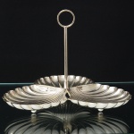 Elegante petisqueira confeccionada em metal espessurado à prata, dotada de três recipientes moldados em forma de concha, apoiados sobre três pés esféricos, interior ligeiramente côncavo, repuxado com sulcos longitudinais, borda recortada em ondulações regulares; haste central tubular encimada por alça circular. Medidas: Alt.: 14,5 x Diam.: 17 cm.