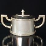 FRACALANZA. Elegante açucareiro confeccionado em metal espessurado à prata, corpo cônico decorado nas bordas com fileira de frisos e fitas cruzadas, padrão "croisé", obtido através da técnica do cinzelado, alças laterais curvada de topo retilíneo, tampa encimada por puxador ovoide. Marca da manufatura puncionada ao fundo. Med.: Alt. 10 x Comp. 19 x Larg. 10 cm.