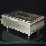 Elegante caixa porta-joias confeccionada em metal espessurado à prata, formato retangular, tampa dotada de abertura basculante, interior revestido em tecido de tom vermelho, superfície decorada com motivo vegetalista centralizando reserva para inserir marca possessória, puxador em aba recortada, assente sobre quatro pés esféricos. Medidas: Alt.: 7 x Comp.: 16 x Larg.: 11 cm.