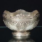 Belíssima fruteira confeccionada em metal espessurado à prata, bojo hemisférico,  ornamentado com painéis verticais delimitados por frisos, preenchidos por elementos fitomórficos e volutas em relevo.borda decorada em relevo com sequência de mascarões femininos alados intercalados por volutas, base circular escalonada, circundada por friso canelado. Execução por técnicas de repuxo, cinzelagem e fundição. Estilo Neoclássico. Medidas: Alt.: 7,5 x Diam>; 13 cm.