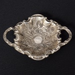 Pequena travessa porta-aliança confeccionada em metal espessurado à prata, formato oval, centro ligeiramente rebaixado, lavrado com flores e folhagens, borda recortada e relevada com motivo rocaille, alças laterais e pés integrados. Medidas: Alt.: 2,5 x Comp.: 9,5 x Larg.: 7 cm.