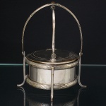 Elegante queijeira confeccionada em metal espessurado à prata, formato circular, bordas cinzeladas com fileiram de frisos e fitas cruzadas, assentado sobre quatro pés curvos, tampa com abertura basculante, alça em arco. Apresenta desgaste no banho de prata. Medidas: