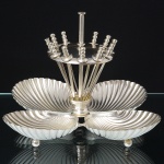 Elegante petisqueira confeccionada em metal espessurado à prata, composto por base central com haste vertical e disco superior perfurado, contendo suportes para espetos removíveis em forma de florete, circundado por quatro recipientes em forma de concha, interior côncavo, repuxado com sulcos e borda recortada em arcos regulares. Apresneta perde de um espeto. Medidas: Alt.: 14 x Comp.: 17 x Larg.: 17 cm.