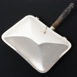 WOLFF. Cata migalhas em metal espessurado à prata, formato retangular de quinas arredondadas, puxador em forma de folhagem curvada, tampa com abertura basculante, cabo em madeira torneada. Med.: Alt. 8 x Comp. 19 x Larg. 25 cm.