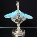 Fruteira vitoriana de pé alto em metal espessurado à prata, receptáculo hemisférico em opalina, borda drapeada, internamentente de tonalidade azul turquesa, verso na cor branco leitoso; alça apresenta decoração relevada com arabescos, enrolamentos vegetalistas e cartelas ornamentadas em estilo neorrococó; nas laterais, efígies femininas em perfil adornam as fixações, representando cabeças aladas de influência clássica, integradas a volutas simétricas, base abaulada cercada por faixa contendo sucessão de folhagens em relevo. Marca da manufatura puncionada na borda da base. Med.: Alt. 26 x Comp. 21,5 x Larg. 22 cm.