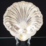 Old English Silver & Co., Sheffield - Inglaterra. Petisqueira em metal inglês espessurado a prata, ricamente moldado com forma de concha, dotado de recipiente para molho, borda recortada em arcos. Marca da manufatura ao fundo. Med.: Alt. 4 x Comp. 28 x Larg. 29,5 cm.