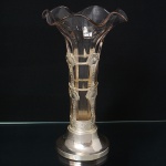 Vaso em vidro com montures em prata, corpo tubular em vidro transparente, com boca moldada em ondulações. Suporte confeccionado em metal espessurado à prata, composto por armação vertical e elementos ovais interligados por hastes, formando estrutura vazada de reforço. Base circular  escalonada, com assentamento do vidro por encaixe. Execução por técnicas de sopro do vidro, fundição, conformação e soldagem do metal. Medidas: Alt.: 30 x Diam.: 16,5 cm.