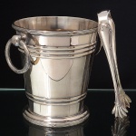 FRACALANZA. Elegante geleira confeccionada em metal espessurado à prata, formato aproximadamente obcônico, assente sobre base circular, bordas relevadas com frisos intercalados por fitas cruzadas, obtidos através da técnica do cinzelado, ornatoreplicado nas alças laterais em argolas. Marca da manufatura puncionada ao fundo. Acompanha um(01) pegador de gelo com pegas em forma de garra. Meds.: Geleira: Alt. 13 x Diam. 14,5 cm; Pegador: Comp. 15,5 cm.