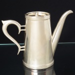 MAGNA PRATA 90. Bule confeccionado em metal espessurado à prata, bojo obcônico com bodas superior e inferior relevadas, tampa circular com puxador torneado, bico vertdor tubular de ponta curvada e recortada, alça lateral em voluta. Marca da manufatura ao fundo. Medidas: Alt.: 125 x Comp.: 16 x Larg.: 7,5 cm. Apresenta marcas de uso.