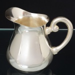 WOLFF. Elegante leiteira confeccionada em metal espessurado à prata, bojo piriforme com alça lateral em voluta, borda ondulada e vertedouro revirado para fora. Marca da manufatura puncionada ao fundo. Medidas: Alt.: 10 x Comp.: 11,5 x Larg.: 9 cm.