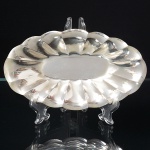 EBERLE. Pequena travessa oblonga confeccionada em metal espessurado à prata, centro rebaixado, orla repuxada com sucessão de sulcos verticais, borda recortada e revirada para fora. Marca da manufatura no verso. Medidas: Alt.: 2,5 x Comp.: 18 x Larg.: 10,5 cm.