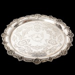 Salva confeccionada em metal espessurado à prata, formato circular, centro ricamente lavrado com composição de motivo fitomorfo, padrão replicado ao redor da borda, assente sobre três pés traingulares relevados com volutas. Medidas: Alt.: 3,5 x Diam.: 30 cm.