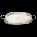 Belíssima bandeja oblonga confeccionada em metal espessurado à prata, centro ligeiramente rebaixado, lavrado com composição de motivo vegetalista, quinas relevadas com flores e folhas de acanto, intercaladas por ondulações que percorrem a borda, padrão decorativo replicado nas alas laterais. Medidas: Comp.: 50 cm(de uma alça a outra) x Larg.: 26,5 cm.