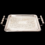 BELPRATA. Elegante bandeja confeccionada em metal espessurado à prata, formato retangular de quinas arredondadas, borda percorrida por contorno relevado, centro ligeiramente rebaixado, ricamente lavrado com composição de motivoi vegetalista, alças laterais fusiformes, relevada com folhagens, apoia sobre hastes curvadas. Medidas: Alt.: 4,5 x Comp.: 40 x Larg.: 25 cm.