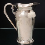 EBERLE. Elegante jarra confeccionada em metal espessurado à prata, bojo balaústre ricamente lavrada com elementos vegetalistas, assente sobre base circular repuxada em degraus, asa lateral em volutas, boda ondulada e revirada para fora, interior dotado de barra retentora de gelo. Marca da manufatura no verso. Med.: Alt. 21,5 x Comp. 20 x Larg. 13 cm.