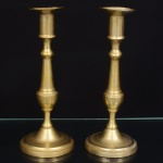 Par de castiçais em metal dourado, corpo torneado, composto por bocal cilíndrico com borda ligeiramente aberta, coluna vertical segmentada por nó central de perfil ovóide, trecho inferior afilado e base circular escalonada, de perfil convexo. Medidas: Alt.: x Diam.: