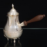 REBOUÇAS PRATA 90. Chocolateira confeccionada em metal espessurado à prata, estilo inglês; bojo piriforme decorado com sucessão de gomos verticais na parte inferior, obtidos através da técnica do repuxado, tampa campaniforme encimada por puxador em pináculo, dotada de abertura através de movimento basculante, assentado sobre três pés de espátula curvada sob concha, alça lateral executada em madeira torneada. Marca da manufatura ao fundo. Medidas: Alt.: 18,5 x Comp.: 20 x Larg.: 10 cm. Apresneta desgaste no banho de prata localizado ao redor da borda.