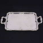 MERIDIONAL INOX. Bandeja confeccionada em aço inoxidável, formato retangular de quinas arredondadas, centro ligeiramente rebaixado, borda repuxada com volutas e elementos conchiformes, padrão replicado nas alças laterais. Etiqueta da manufatura no verso. Medidas: Comp.: 53 x Larg.: 31 cm.