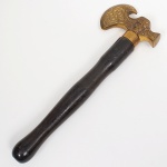 Saca-rolhas em forma de machado, composto por cabeça em metal amarelo, com lâmina curva semicircular configurada como machado estilizado, ornamentada por gravação em relevo de volutas. Lâmina destacável por rosqueamento, que, ao ser removida, revela saca-rolhas metálico helicoidal integrado ao corpo da peça. Encaixe da cabeça ao cabo por sistema de rosca. Cabo em madeira, torneado, de seção circular, com empunhadura alongada e virola metálica lisa na extremidade superior. Medida: Comp.: 21,5 cm.