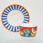 Fábrica de Porcelana da Vista Alegre, ílhavo - Portugal(Fundada em 1824 por José Ferreira Basto). Xícara para café da manhã em faiança portuguesa da década de 1980/90, padrão 'Market Place'; esmaltação branca, decoração composta por faixa contínua contendo  frutas e flores, incluindo melancia e arranjo floral, delimitada por filetes lineares, aplicadas em estampagem policromada, pires circundado com motivo radial estilizado em azul e branco, borda contornada por frisos nas tonalidades azul e vernelho. Marca da manufatura no verso. Medidas: Xícara: Alt.: 9,5 x Comp.: 15 x Larg.: 11,5 cm; Pires: Diam.: 20,5 cm.