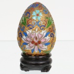 Ovo decorativo em esmalte cloisonné,  corpo inteiriço, decorado em policromia por compartimentação em fios metálicos formando composição floral central estilizada, envolvida por volutas e elementos fitomórficos. Base  em madeira entalhada, de formato circular com pés baixos. Medidas: