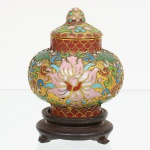 Potiche em esmalte cloisonné, corpo globular com bojo expandido, gargalo curto e tampa em cúpula com botão superior. Decoração policromada executada por compartimentação em fios metálicos, formando composição floral central estilizada, envolvida por volutas e padrões geométricos. Gargalo e tampa decorados com faixas ornamentais repetitivas. Base apoiada sobre suporte em madeira entalhada, de formato circular escalonado, com pés baixos. Medidas: