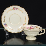 KPM - Krister Porzellan-Manufaktur, Waldenburg - Alemanha(Fundada em 1831 por Karl Kristen). Excepcional chávena para chá em porcelana de manufatura alemã, fabricada na polônia entre anos de 1930-1945, modelo Friederike, citado na Enciclopédia de Porcelana da Silésia Volume I. Série "Royal Ivory". Executado ao estilo renascentista. Esmaltagem vítrea de tom marfim, modelada com elementos fitomorfos em relevo, decorada com flores soltas em estampagem policromada, e generosos arremates de frisos em douraudura, chávena de bojo campaniforme, assente sobre base alta de formato circular e alça lateral em voluta, padrão decorativo replicado no pires e no prato. Marca da manufatura no verso. Meds.: Chávena: Alt. 6 x Comp. 12 x Larg. 10 cm. Pires: Diam. 15 cm.