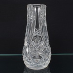 Vaso executado em espesso cristal, diáfano e incolor, formato baalústre, de perfil vertical, com boca circular levemente evertida e base circular plana; corpo decorado por painéis facetados em alto-relevo, organizados em composição geométrica simétrica, alternando estrelas radiadas, losangos facetados e frisos verticais sinuosos que acompanham o eixo do vaso, região central marcada por medalhão estrelado, integrado ao conjunto decorativo, fundo com estrela radial moldada, circundada por anel circular em relevo. Apresenta pequenos desgastes ao redor da borda superior, e marcas de uso distribuídas no corpo. Medida: Alt.: 22 x Diam.: 10 cm.