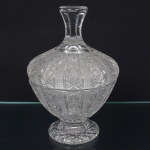 Bombonnière executada em cristal europeu, diáfano e incolor, corpo abaulado com tampa correspondente, formato ovóide, assentada sobre pé circular elevado, decorados por lapidação geométrica em alto-relevo, organizada em painéis verticais facetados, alternando campos de pequenos losangos em treliça, rosetas circulares lapidadas e frisos verticais polidos, distribuídos de forma radial. Transição corpo/tampa marcada por linha de encaixe contínua. Base com estrela radial lapidada, circundada por anel liso. Tampa com pegador integrado em formato cilíndrico afunilado, decorado por anelamentos horizontais regulares. Perfeito estado, sem bicados ou trincas. Medida: Alt.: 23 x Diam.: 15 cm.