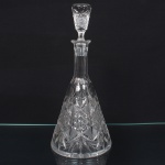 Exuberante garrafa executada em cristal europeu, diáfano e incolor, bojo aproximadamente cônico profusamente talhado com padrão de folhas estilizadas, estrelas e formas geométricas em relevo, organizado em painéis verticais facetados, base circular, fundo com lapidação radial em estrela, com raios  partindo do centro em disposição simétrica, gargalo facetado, tampa obcônico decorada com o mesmo padrão. Medida: Alt.: 32,5 x Diam.: 14,5 cm.