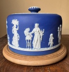 Wedgwood , porta-bolo em Jasper azul escuro e branco com decoração de vestais em estilo neoclàssico, marcado na base em baixo relevo Wedgwood, base em madeira, 16x25 cm . Em 1738 começaram as escavacões de Pompéia e Herculano próximo de Nápoles na Itália criando a partir dessa época na Europa um novo interesse ao estilo neoclássico. Em 1759 Josiah Wedgwood fundou uma manufatura de porcelana inspirada em motivos da antiguidade clássica, tornando sua empresa consagrada mundialmente. As peças eram realizadas com um tipo de material ao qual foi dado no nome Jasper.