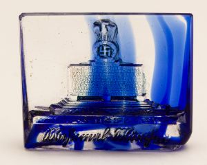 Militaria, Alemanha II Guerra. Proveniente de importante coleção adquirida na Bélgica - Feira de Militaria de Ciney. O partido nazista promoveu uma grande campanha de arrecadação de fundos antes e durante a guerra, com a venda de itens colecionáveis, pins, pendentes, tinnies, badges etc. Sempre colecionáveis voltada a novas compras. Oportunidade para colecionador avançado. **Item não promove ou glorifica violência ou intolerância racial. Está à venda apenas para fins de preservação da memória histórica. A Organização do leilão não faz apologia a quaisquer movimentos políticos ou ideológicos, e repudia qualquer ideologia de cunho racista. Em vidro. 33x26 mm. Monumento Nazista com Águia sobre Cruz Gamada - Rara.