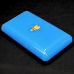 RAINHA VICTORIA (1819-1921) GRANDE PESO DE PAPEL EM BRISTOL GLASS (VIDRO OPALINO PRODUZIDO NA INGLATERRA ENTRE OS SECULOS XVII E XIX). EXCECUTADO COMO PEÇA COMEMORATIVA DA COROAÇÃO DA MONARCA EM 20 DE JUNHO DE 1837. APLICAÇÃO DA EFIGIE DA RAINHA EM BRONZE ORMOLU SOBRE GRANDE PLACA EM VIDRO OPALINO AZUL. INFLATERRA, 1837. 17 X 10 CMNOTA: Em 1651, os mestres do ferro na Floresta de Dean, enfrentando problemas na fabricação do poste onde fundiam o minério, chamaram um engenhoso mestre vidreiro, Edward Dagney, um italiano que então vivia em Bristol. Dagney provavelmente era um dos membros da família Dagnia, que não continuou trabalhando em Bristol, e os filhos de Edward, Onesiphorus, mudaram-se para Tyneside.Há referências anteriores a vidreiros em Bristol, no final do século XIII e início do século XIV , por exemplo, mas sem nenhuma indicação precisa da natureza de sua profissão. Vidraceiros aparecem nos livros de aprendizes do século XVI , o que não é surpreendente, visto que a demanda por vidro para janelas para uso eclesiástico e, quando o padrão de vida melhorou, para fins domésticos, foi um dos principais estímulos para a indústria vidreira local em seus primórdios. Foi na virada do século XVII , no entanto, que surgiram evidências definitivas da dimensão e da natureza da indústria vidreira de Bristol . Em 1969, John Houghton, em uma de suas cartas sobre agricultura e comércio, listou as fábricas de vidro na Inglaterra e no País de Gales. Ele registrou que "em Bristol e arredores" havia cinco fábricas de garrafas, uma de garrafas e vidro para janelas, e três de vidro de sílex e vidro comum. Era, fora de Londres, a maior concentração de fábricas de vidro.   Um dos fatores que contribuíram para o crescimento bem-sucedido do comércio em Bristol, e consequentemente da indústria vidreira, foi a estabilidade política do condado sob o governo de Walpole. A guerra com a França terminou em 1713, e as políticas financeiras e econômicas subsequentes do governo impulsionaram o crescimento industrial e comercial. Contudo, essas políticas não foram isentas de problemas. Houve um ataque à evasão de direitos alfandegários, e a proibição da importação de vinho em garrafas foi uma das formas pelas quais a indústria vidreira foi afetada. Além disso, quando se tentou introduzir armazéns alfandegados para vinho e tabaco, as propostas encontraram forte oposição e tiveram que ser abandonadas. Ademais, a tentativa de conter o aumento do alcoolismo por meio de impostos proibitivos foi abertamente desafiada, e embora o ditado "bêbado por um centavo, completamente bêbado por dois centavos" possa ter estimulado a venda de garrafas de vidro, não contribuiu em nada para aumentar a confiança do público em uma tributação eficaz. Por fim, os anos de paz chegaram ao fim, e mais uma vez o governo se viu diante da necessidade de arrecadar fundos para a guerra. Em 1745, a indústria do vidro se viu novamente sobrecarregada por um imposto que, meio século antes, havia eliminado com sucesso, e foram necessários cem anos para que o fardo fosse removido.A fabricação de vidro no século XVIII permaneceu praticamente inalterada por quase dois mil anos, e seus métodos são semelhantes aos usados hoje em dia na produção artesanal de vidro. Sempre cativou a imaginação, e aqueles que hoje fazem fila para ver o vidro sendo feito dessa maneira eram iguais aos seus antepassados em Bristol. Shiercliff, em seu Guia de Bristol e Hotwell de 1789, escreveu: "...e para aqueles que nunca viram a maneira de trabalhar esse material, pode ser um entretenimento agradável assistir ao processo... os visitantes nunca são impedidos de ver as pessoas trabalhando, mediante uma pequena gorjeta dada aos empregados". Os vidreiros sempre se orgulharam de suas habilidades. Tão confiantes, aliás, que estão dispostos a tentar fazer qualquer coisa em vidro. Quando o Príncipe e a Princesa de Gales visitaram Bristol em 1738, o relatório dizia o seguinte:'A companhia dos Homens de Vidro foi a primeira a marchar, vestidos com camisas brancas holandesas, alguns com espadas, outros com coroas e cetros de vidro nas mãos.'Essas peças, conhecidas como "friggers", eram geralmente feitas durante o intervalo ou no final do dia, com os restos de vidro do fundo da panela, e era uma das maneiras pelas quais o vidreiro aprimorava suas habilidades e, frequentemente, seu dinheiro, já que eram vendidas fora da fábrica como lembranças. Nem todos os comentaristas viam os vidreiros sob uma luz tão romântica. Patty More, ao visitar as fábricas de vidro de Nailsea com sua irmã no final do século XVIII, foi pouco elogiosa ao escrever:'O trabalho em uma fábrica de vidro é irregular, incerto se é dia ou noite; não só infringe o descanso do homem, como também invade constantemente os privilégios do sábado. Os salários são altos, a comida e a bebida são luxuosas  o corpo mal coberto, mas alimentado com iguarias de gosto duvidoso. Os altos edifícios das fábricas de vidro enfileirados diante das portas dessas casas  as grandes fornalhas rugindo  os palavrões, a comida e a bebida desses seres seminus e de aparência sombria, conferiam ao local um aspecto infernal e horrível.'     Embora o vidro para janelas, e em particular para garrafas, constituísse a base da indústria vidreira de Bristol, sua reputação foi estabelecida pelos fabricantes de vidro de sílex. Em 1675, foi introduzido o vidro com alto teor de óxido de chumbo, o chamado "vidro de chumbo". Devido à facilidade com que podia ser trabalhado, à sua transparência e à sua qualidade, esse vidro logo começou a substituir o vidro de sílex convencional à base de soda. No final do século XVII, a maioria, senão todas, as fábricas de vidro de Bristol que produziam artigos domésticos, como copos, já haviam adotado o vidro de chumbo.Foi, contudo, na segunda metade do século XVIII que o nome de Bristol entrou para a história do vidro. Foi durante esse período que os vidros coloridos se popularizaram, particularmente o vidro azul, posteriormente associado à expressão "azul Bristol". O vidro azul não se restringiu a esse período, nem era exclusivo de Bristol, visto que o uso do cobalto como meio de obtenção dessa cor era conhecido pelos fabricantes de vidro há séculos. Entretanto, foi o cobalto proveniente de uma fonte específica na Saxônia que, no século XVIII , foi considerado o responsável pela qualidade de cor insuperável. Durante o terceiro quarto do século, o fornecimento britânico de cobalto dessa fonte parece ter estado nas mãos de William Cookworthy, um fabricante de porcelana e comerciante de matérias-primas do oeste da Inglaterra, que havia construído consideráveis interesses em Bristol. Como Cookworthy não impunha restrições quanto a quem podia comprar de seu estoque de cobalto, este estava prontamente disponível para fabricantes de vidro em todo o país. Assim, o termo "azul de Bristol" pode igualmente ter surgido do fato de Bristol ser o local onde esse corante era adquirido. Um desenvolvimento posterior na segunda metade do século XVIII foi o do vidro branco opaco, que se assemelhava à porcelana, mas custava muito menos. Amplamente produzido em todo o país, a qualidade do produto foi aprimorada por decoradores que pintavam flores, pássaros, motivos chineses e outros na superfície. O mais notável entre eles foi Michael Edkins, que iniciou sua carreira na fábrica de cerâmica de Delft em Redcliff, Bristol. Michael Edkins abriu seu próprio negócio por volta de 1760 e decorou peças para a fábrica de vidro Redcliff Backs, para a fábrica de vidro de curta duração de William Dunbar and Co. em Chepstow e para o comerciante de vidro de Bristol, Lazarus Jacobs. Devido à pequena quantidade produzida na época, o vidro branco opaco não foi incluído na lei de impostos de 1745. Essa omissão foi corrigida em 1778, quando o produto foi incluído na lista juntamente com o vidro de sílex, cujo preço foi duplicado naquele ano, acabando efetivamente com o comércio de vidro branco opaco.     Qualquer esperança de que o imposto sobre o consumo fosse rapidamente retirado foi frustrada quando, em 1756, foi declarada guerra à França. Pior ainda estava por vir, devido à política do governo em relação às colônias americanas. O atrito entre os dois lados levou, eventualmente, a um acordo sem importância por parte de algumas colônias. Nada poderia ter atingido os comerciantes de Bristol com mais força, e em 1775, os Mercadores Aventureiros enviaram uma petição ao Parlamento. Eles citaram o efeito que certos impostos anteriores haviam tido sobre o comércio de "vidro, papel e outros artigos". Esse comércio havia se recuperado desde a substituição desses impostos, mas outras medidas estavam causando inquietação. Concluíram com uma referência aos "milhares de miseráveis súditos que, com a paralisação total das exportações para a América, serão demitidos de suas fábricas por falta de emprego e ficarão em grande situação de pobreza".Contudo, apesar da imposição do imposto sobre o consumo, a produção de vidro no Reino Unido aumentou gradualmente até que, no início da Guerra da Independência Americana, em 1775, era cerca de 70% superior à de 1745, quando o imposto foi introduzido. Mesmo assim, em três anos a produção caiu um terço e só foi possível recuperar o nível anterior dez anos depois. Os fabricantes de vidro de Bristol, dependentes das colônias americanas para grande parte do seu comércio, podem ter sofrido ainda mais, visto que foi nesta década que se iniciou o declínio de Bristol como centro de fabricação de vidro. Diversas fábricas de vidro foram colocadas à venda, à medida que alguns dos comerciantes e fabricantes de vidro mais conhecidos começaram a abandonar o ramo. Desde a onda de investimentos do início do século, apenas mais duas fábricas de vidro foram construídas: uma em Crews Hole e a outra na continuação da Red Lane, que mais tarde se tornaria a Prewitt Street. Agora, com a indústria em declínio, uma fábrica de vidro foi construída no local da Phoenix Inn, na Portwall Lane. O homem por trás dessa empreitada era Jacob Wilcox Ricketts, possivelmente o mais implacável dos empresários da indústria vidreira de Bristol, cujos outros interesses incluíam tabaco e cerveja. Nos vinte anos seguintes, Ricketts reuniu todas as fábricas de vidro de sílex de Bristol, reabriu uma fábrica de vidro para garrafas e obteve um lucro considerável. Tudo isso enquanto a indústria vidreira em Bristol encolhia para cerca de um quarto do seu tamanho anterior.Em 1820, a produção de vidro de sílex e garrafas no Reino Unido caiu drasticamente. A bem-sucedida fábrica de vidro Phoenix começou a acumular grandes prejuízos e nunca mais recuperou sua lucratividade anterior, enquanto a empresa de decoração de vidro de Lazarus Jacobs, descrita em um anúncio como "fabricante de vidro para Sua Majestade", para a qual Michael Edkins trabalhara desde 1763, faliu. Na ocasião anterior em que um imposto sobre o consumo havia sido imposto, os fabricantes de vidro de Bristol manifestaram suas objeções, mas desta vez foram menos veementes. Quando o governo instaurou uma investigação sobre seus efeitos, a fábrica de garrafas de Limekiln Lane estava prestes a fechar, a família Rickets parecia ter perdido o entusiasmo, e coube a William Powell, proprietário da fábrica de vidro Hoopers na Avon Street, apresentar o ponto de vista de Bristol.O relatório dos Comissários, quando publicado em 1835, foi particularmente revelador. Listava 106 fábricas de vidro na Inglaterra e 10 na Escócia. A maioria das fábricas de vidro estava agora nas Midlands e no norte, sendo o nordeste, com 41 fábricas, a área predominante. Bristol, com quatro fábricas, Nailsea, com duas, e Londres, com três, eram as únicas áreas no sul onde a fabricação de vidro continuava. Pouco menos de 10% de todas as garrafas, menos de 3% do vidro de sílex e nenhum vidro de coroa eram agora fabricados em Bristol. A fabricação de vidro havia se deslocado inexoravelmente para as regiões onde o carvão era mais facilmente obtido e onde as crescentes cidades industriais tinham melhores meios de comunicação com seus mercados.Por isso a produção de Bristol é extremamente rara e atinge preços altos no mercado. 