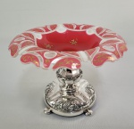 MARCUS & CO - REQUINTADA EPERGNE EM MAGNIFICO VIDRO ARTISTICO DA BOHEMIA SOB O IMPÉRIO AUSTRO HUNGARO. SUNTUOSO TRABALHO EM OVERLAY NAS CORES BRANCA, ROSE E TRANSLUCIDO REMATADOS COM ROCAILLES EM ESMALTES REALÇADOS COM OURO. BASE EM PRATA DE LEI COM CONTRASTES EUROPEUS E MARCA DE IMPORTAÇÃO DA PRESTIGIADA JOALHERIA MARCUS & CO DE NOVA YORK. PERIODO ÁUREO DESTA IMPORTANTE JOALHERIA NA VIRADA DO SEC. XIX/XX. 23 X 17 CM . 870 G NOTA: A Marcus & Co. era uma joalheria americana, sediada na cidade de Nova Iorque, famosa por suas peças de alta qualidade nos estilos Art Nouveau e Art Déco.Herman Marcus, um joalheiro alemão, fundou a empresa com seu filho William em 1892. Antes de se mudar para Nova York em 1850, Herman Marcus trabalhou para Ellemeyer , uma prestigiada joalheria da corte em Dresden. Foi na Europa que ele descobriu técnicas de esmaltação que não eram comuns nos EUA naquela época. Quando se estabeleceu nos EUA, foi seu conhecimento e savoir-faire que lhe garantiram empregos na Ball, Black & Co. (mais tarde Black, Star & Frost) e na Tiffany & Co. As peças de prata de lei  e vidro da Marcus & Co. são itens de colecionador altamente valorizados, produzidos principalmente entre o final do século XIX e o início do século XX. A empresa era famosa por sua maestria em estilos como Art Nouveau e Art Deco, frequentemente incorporando técnicas complexas como o esmalte plique-à-jour para criar efeitos semelhantes a vitrais. A Marcus & Co. possuía um departamento especializado em prataria que colaborava com fabricantes renomados para criar peças luxuosas para a elite de Nova York.  As peças autênticas do período áureo (18921920) apresentam designs orgânicos, formas assimétricas e detalhes de esmalte aplicados em superfícies curvas, o que era uma especialidade da casa.