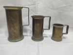 Conjunto com três canecas medidoras antigas em bronze, de tamanhos graduais, com alças laterais fixas, bases circulares e pátina original do tempo, apresentando marcas de uso e pequena mossa compatíveis com a idade, peças decorativas e colecionáveis. Medindo o maior 19cm de altura.