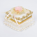 LOUÇA, uma (1) caixa com tampa, quadrangular, abertura basculante, monture em metal dourado, tampa relevada em elementos vegetalistas na própria modelagem, tonalidade branca, apliques de barrado e flores em tecido, sem marca de manufatura aparente, medindo 7,5 x 7,7 x 5 cm, sinais de uso, vide foto.