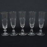 CRISTAL E VIDRO, cinco (5) flutes de pé alto, incolores e translúcidos, dois modelos distintos: 4 com bojoobcônico, metade inferior lapidação dedão, fuste no formato de colunetas sobrepostas, base em bolacha, medindo 17 cmaltura; 1 com bojo obcônico, metade inferior lapidação dedão, fuste no formato de balaustre, base em bolacha, medindo17,5 cm altura, todos com sinais de uso, vide foto.