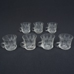 VIDRO, sete (7) canecas pequenas, incolores e translúcidas, bojo lapidado em sulcos e fecho de trigo, fundo em dália, quatro deles com bicados na borda, medindo 7 cm altura, sinais de uso, vide foto.