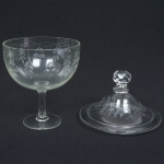 VIDRO, uma (1) compoteira de pé alto, incolor e translúcida com tampa adaptada, bojo com singela lapidação vegetalista, fuste quase cilíndrico, base em bolacha, tampa com ornamentação floral pirografada, pega facetada com sinais de que foi relapidada, medindo 25 cm altura, sinais de uso, vide foto.