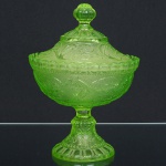 VIDRO, uma (1) antiga compoteira de pé alto com tampa, translúcida, tonalidade verde limão, ornamentação modelada em relevo com dragões, perolados, caneluras e elementos vegetalistas, medindo 21 cm altura, sinais de uso, vide foto.